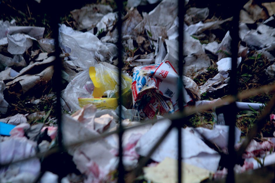 A close-up view of a pile of discarded rubbish behind a black metal fence, containing crumpled paper, plastic packaging, and printed cardboard with red and blue graphics. The debris is scattered on the ground, with some items partially obscured by the fence bars. The rubbish includes torn and flattened materials with varied textures such as glossy plastic and matte paper, in colors like white, yellow, red, and blue. The background shows soil and fallen leaves, indicating an outdoor environment, possibly a waste disposal or fly-tipping site. The lighting is natural, casting soft shadows on the scattered refuse. This scene illustrates the type of waste that may be subject to independent collection or private disposal before it is cleared by rubbish removal services like those offered by companies such as rubbishclearancekentishtown.com, emphasizing the importance of proper waste handling and adhering to disposal rules.