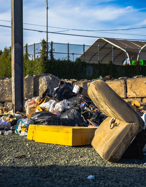 A pile of mixed rubbish consisting of black plastic garbage bags, cardboard boxes, and discarded household items, including a worn and dirty seat cushion leaning against the heap. The waste is situated on an asphalt surface near a stone wall constructed from uneven, rough-textured rocks. In the background, there is a black metal fence with vertical bars, and beyond that, a tennis court with a white, semi-transparent canopy structure. To the left, a tall metal utility pole with overhead electrical wires extends into the sky, which is clear with some scattered clouds, illuminated by bright sunlight. The scene suggests an outdoor rubbish accumulation area possibly awaiting independent collection or private disposal, typical of non-local-authority waste handling contexts. The rubbish appears haphazardly piled, indicating it has been deliberately left there without proper clearance, reflecting practices associated with non-standard waste removal services or fly-tipping avoidance.