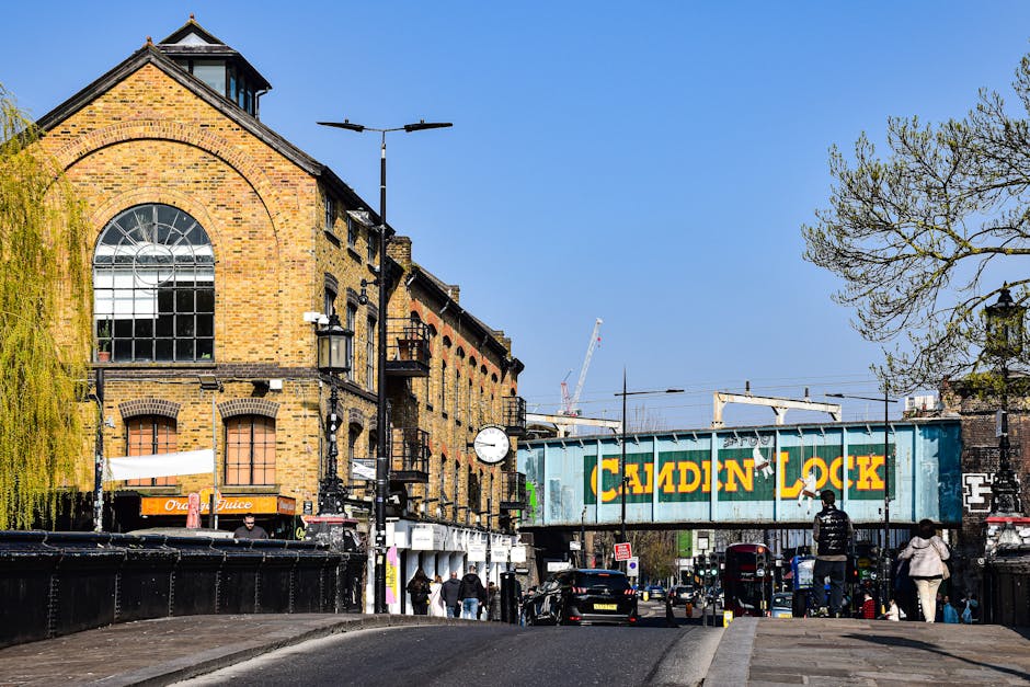 A street scene in Kentish Town featuring a large, weathered blue freight train carriage with the words 'Camden Lock' painted in yellow on its side, positioned across the road beneath an elevated railway track. The surrounding environment includes a historic brick building with arched windows on the left, which has a mixture of old brickwork and modern elements like street lamps and a clock mounted on the building facade. The street is populated with pedestrians walking along the pavement, some near a small retail outlet with an orange sign and others crossing the road. Several cars and a bus are visible in the background, along with a few trees on the right side, one of which has sparse, budding branches indicating early spring. The sky is clear and blue, casting natural daylight over the scene, which closely relates to urban waste management and the potential for private rubbish removal services such as those offered by rubbishclearancekentishtown.com, especially in contexts involving on-site clearance or alternative waste handling methods around Camden's iconic areas.