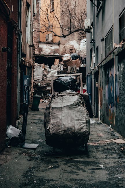 A narrow urban alleyway filled with accumulated waste and debris, featuring a large, weathered black refuse bag in the foreground, which is slightly sagging and appears to contain mixed rubbish. Behind it, there is a cluttered pile of cardboard boxes, plastic bags, and other discarded items stacked against an uneven wall with peeling paint and graffiti. The alley's pavement shows signs of dirt, oil stains, and scattered trash, extending towards a background of leafless trees and brick buildings with a muted, overcast sky overhead. The scene suggests an informal outdoor rubbish collection area, possibly serviced by a private waste removal company such as Rubbish Clearance Kentish Town, emphasizing the need for organized waste disposal to maintain street cleanliness and environmental standards in urban settings. The environment appears neglected, typical of an secluded alley awaiting clearance or scheduled for waste removal services, with the surrounding walls showing signs of age and graffiti, and a slight haze of dust or grime enhancing the urban aesthetic.