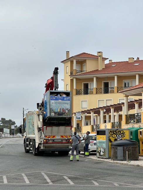 A waste collection vehicle with an extended hydraulic arm is parked on a paved urban street, actively involved in rubbish removal. The truck's rear compartment is partially open, revealing a compartment designed for lifting and emptying large containers. Two workers, dressed in high-visibility trousers and working jackets, are positioned near the back of the vehicle, engaged in managing or guiding the collection process. They are standing next to a green and yellow recycling bin, with graffiti visible on it, and a black trash bin situated on the sidewalk nearby. In the background, a multi-storey residential building with a yellow facade, white window frames, and black wrought iron balcony railings dominates the scene. The building features a red-tiled roof and multiple balconies, some with potted plants, contributing to the residential environment. The sky is overcast, casting diffuse natural light over the scene, which is typical for daytime rubbish collection activities in the area. This image, sourced from rubbishclearancekentishtown.com, illustrates an example of private waste handling involving on-site collection services rather than council rubbish removal, aligning with alternative waste disposal methods for residents and local businesses.