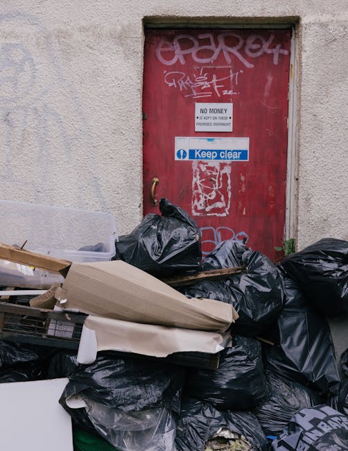 A collection of discarded rubbish including black plastic rubbish bags, flattened cardboard boxes, and miscellaneous waste items piled on a pavement in front of a red metal door, which is covered in graffiti and stickers. The door features a white sign indicating 'NO MONEY' with additional text, and a blue 'Keep clear' notice. The scene is set outside a building with a textured, off-white wall, and the rubbish appears to be a result of a nearby flats' waste clearance, possibly handled through private disposal services like those offered by rubbishclearancekentishtown.com. The environmental context suggests a typical urban alleyway or exterior communal area where waste is temporarily accumulated before collection or removal, reflecting the importance of proper rubbish management and independent waste clearance options in the area.