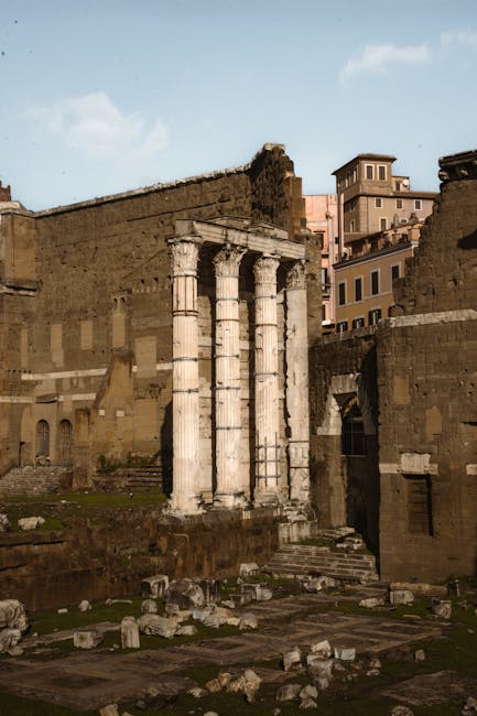The image depicts the remains of ancient Roman columns constructed from white marble, featuring three standing columns with fluted shafts and Corinthian-style capitals. These columns are positioned on a raised stone platform amidst ruins that include scattered fragments and broken stone blocks, possibly part of a collapsed wall or structure, with weathered surfaces and signs of age. In the background, there is a large, partially preserved stone wall made of roughly hewn, beige-colored blocks, showing signs of erosion and damage, with some sections missing or crumbling. Behind the ruins, modern urban buildings of mixed height and design rise against a bright, partly cloudy sky, suggesting a historic site situated within or near a contemporary cityscape. The scene is illuminated by natural daylight, casting soft shadows and highlighting the textures of the ancient stonework and the surrounding environment. The overall setting appears to be an archaeological site engaged in or conducive to private or independent restoration efforts, with the visual emphasis on the historical architecture amid a modern setting, relevant to the themes of alternative waste handling and site clearance by specialist services such as those offered by rubbishclearancekentishtown.com.