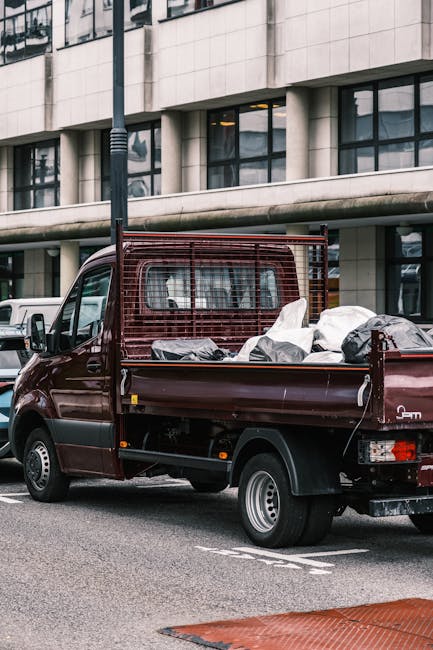 A small burgundy flatbed pickup truck parked on an urban street in front of a modern multi-storey office building with large glass windows and surrounding concrete facade. The truck's bed is filled with various types of waste and rubbish, including large white and black plastic bags, some appearing to contain household or commercial rubbish. A red metal mesh barrier is installed at the back of the bed, possibly to secure the load during transit. The vehicle's rear is close to the curb, and additional parked vehicles are visible in the background. The scene suggests an instance of private waste removal or on-site clearance, characteristic of alternative rubbish collection services, with the presence of waste material indicating recent loading for disposal. Natural daylight illuminates the scene, highlighting the textures of the plastic bags, the metallic surface of the truck, and the contemporary urban setting, consistent with rubbish clearance operations by companies like rubbishclearancekentishtown.com in North West London.