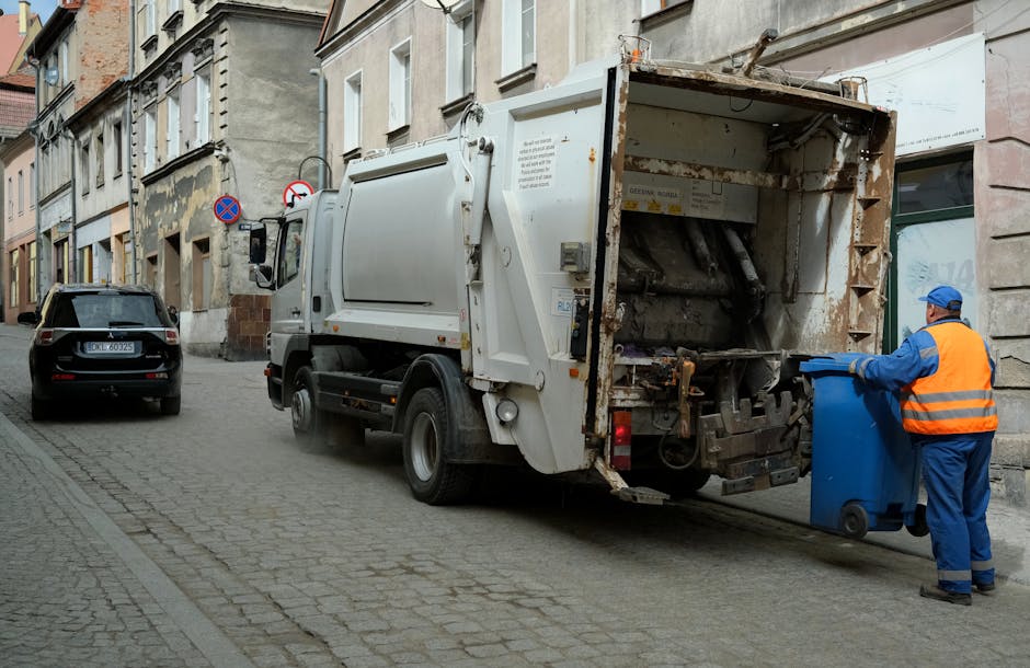A small burgundy flatbed pickup truck parked on an urban street in front of a modern multi-storey office building with large glass windows and surrounding concrete facade. The truck's bed is filled with various types of waste and rubbish, including large white and black plastic bags, some appearing to contain household or commercial rubbish. A red metal mesh barrier is installed at the back of the bed, possibly to secure the load during transit. The vehicle's rear is close to the curb, and additional parked vehicles are visible in the background. The scene suggests an instance of private waste removal or on-site clearance, characteristic of alternative rubbish collection services, with the presence of waste material indicating recent loading for disposal. Natural daylight illuminates the scene, highlighting the textures of the plastic bags, the metallic surface of the truck, and the contemporary urban setting, consistent with rubbish clearance operations by companies like rubbishclearancekentishtown.com in North West London.