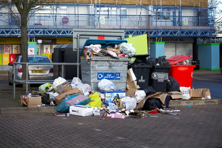 A collection of overflowing rubbish bins and scattered waste on a paved sidewalk in front of a commercial building. The pile includes a large grey mixed paper and card bin with its lid partially open, revealing crumpled newspapers, cardboard, and paper packaging. Surrounding the bins are black, red, and green bin bags, along with loose cardboard boxes, discarded packaging, plastic bottles, and miscellaneous rubbish. The scene is illuminated by natural daylight, with a metal rail in the foreground and a tree trunk on the left side. Behind the waste, there is a storefront with closed roller shutters, bright signage, and a building under construction or refurbishment, indicated by scaffolding and protective netting. The image visually demonstrates the need for efficient rubbish clearance services, such as those provided by [COMPANY_NAME], for managing excess waste through private disposal or on-site clearance efforts in busy urban areas.