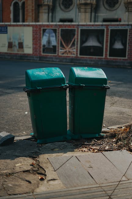 Two green plastic wheelie bins with black lids are positioned side by side on a pavement near the edge of a road. The bins are made of textured, durable plastic with a slight curve on the lids, which are secured with black plastic clips. They are slightly weathered, with minor scuff marks visible on the surface. The bins are placed on a concrete and gravel area that shows some scattered debris, such as dried leaves and small stones, at their base. Behind the bins, there is a wide asphalt road with a slight curve, and across the street, a brick and plaster wall features a series of large advertisements or posters protected by glass, mounted on a building with architectural detailing, including arched windows and ornamental brickwork, indicating an urban setting. The lighting suggests a clear day with natural sunlight casting soft shadows, providing an everyday scene related to local waste collection and private rubbish disposal, which could be managed through services such as those offered by [COMPANY_NAME].